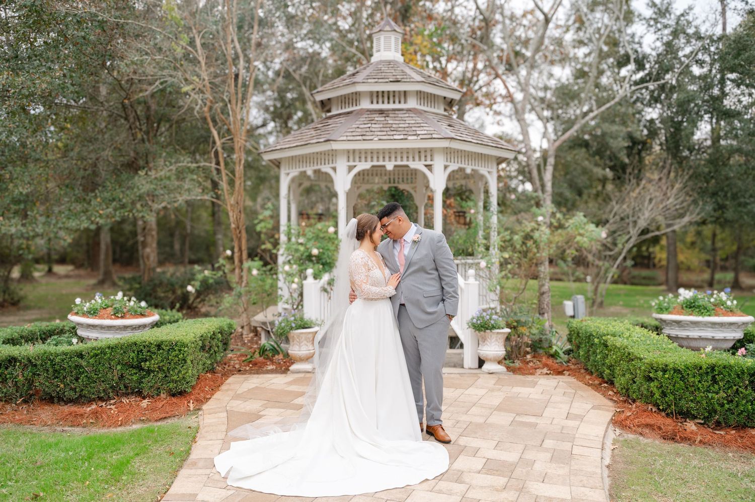 A happy couple poses together on their wedding day, wearing a gray suit and white lace gown with veil.