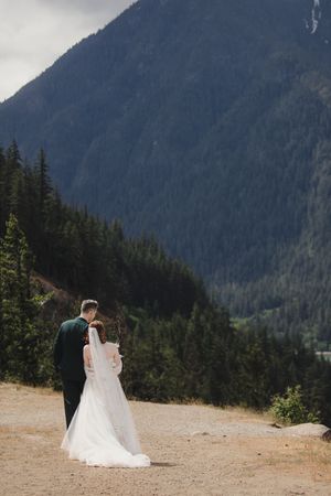 Couple walking along a scenic mountain trail with evergreen forest and misty peaks in the background.