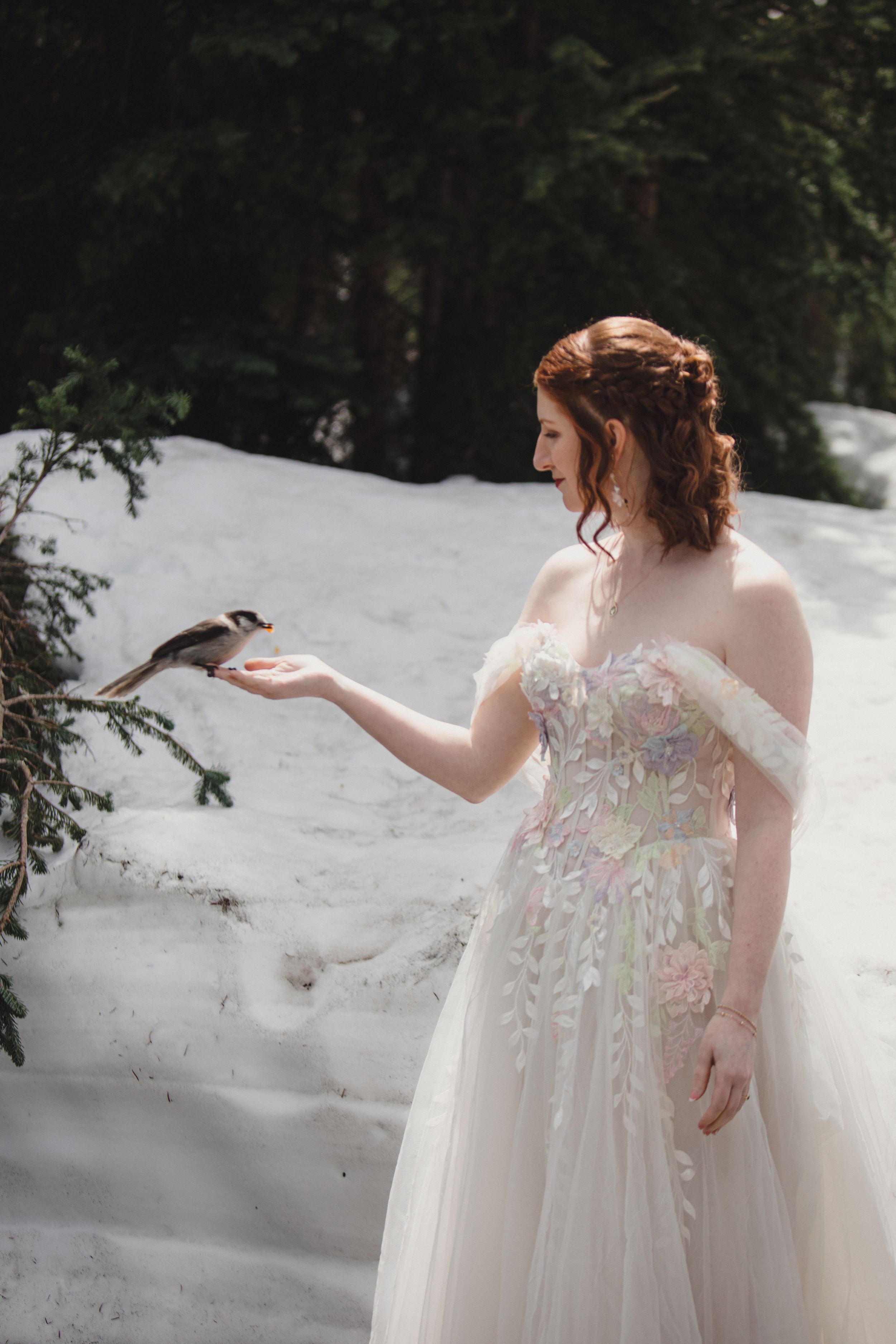 A woman in a flowing white wedding dress walks through fresh snow in a winter wonderland forest setting.