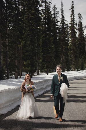 A stylish wedding couple poses playfully on a snowy mountain road lined with evergreen trees.