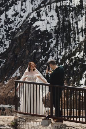 A couple shares a romantic moment at a snowy mountain overlook with snow-covered evergreen trees in the background.