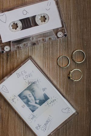 A vintage cassette tape with a handwritten label lies on a wooden surface next to some rings.