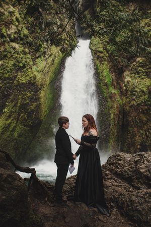 A couple in formal black attire stands together on mossy rocks in front of a majestic waterfall in a lush forest setting.