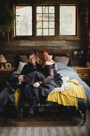 Couple in black clothing share an intimate moment on a cozy bed with yellow blanket by rustic cabin windows.