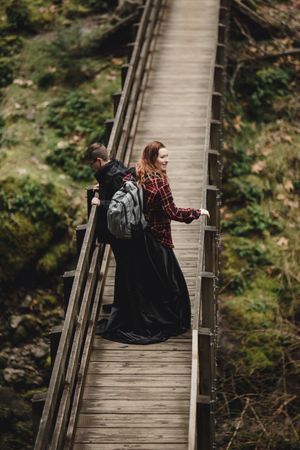 Two people in dark clothing walk along a wooden suspension bridge footpath surrounded by lush greenery.