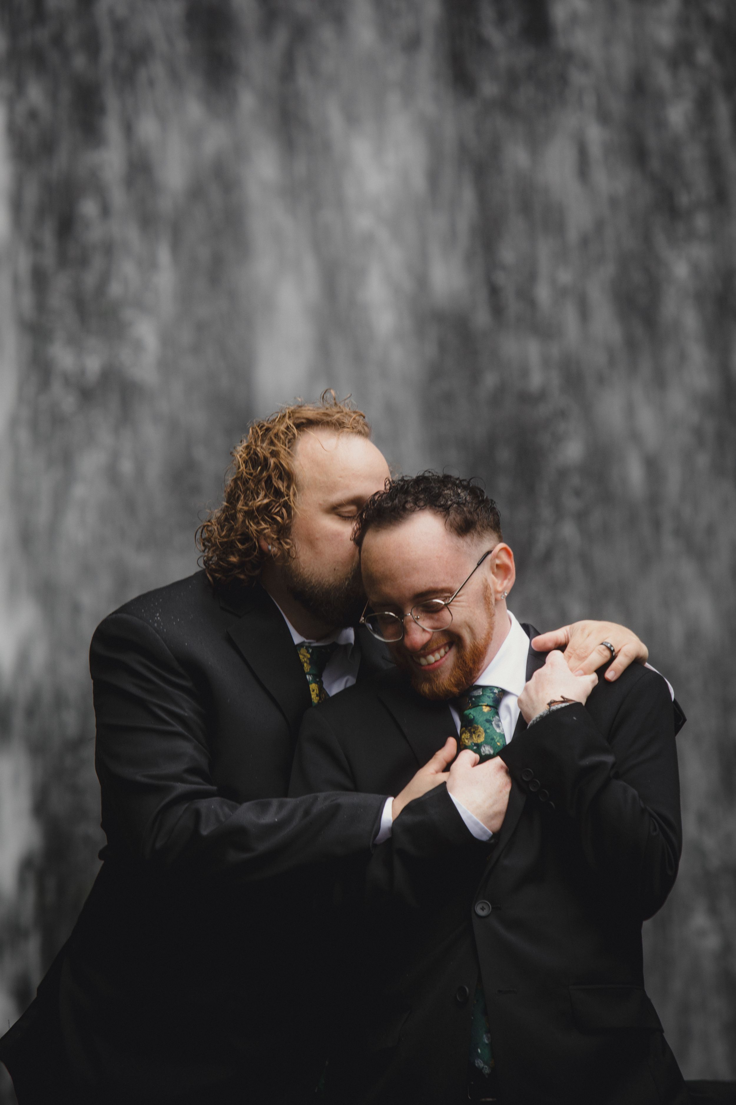 Against a textured grey backdrop, two people in dark suits share emotional laughter in a series of candid moments.