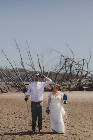 A couple in wedding attire stands on a beach with driftwood, shielding their eyes from the bright sunlight on their big day.