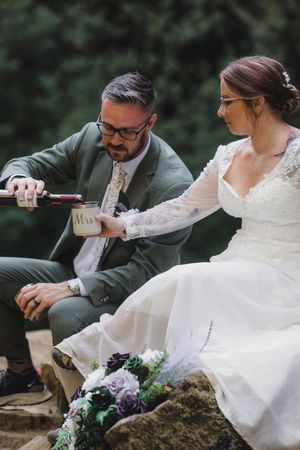Newlywed couple sits on rocks outdoors while pouring drinks together, with bride in lace dress and floral bouquet nearby.