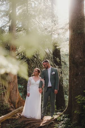 A couple walks together along a forest path on their wedding day surrounded by tall evergreen trees.