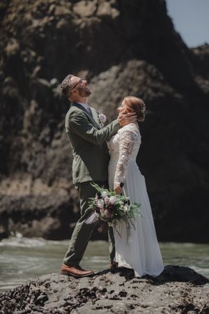 A couple stands on rocky coastal cliffs in wedding attire with the ocean waves crashing behind them.