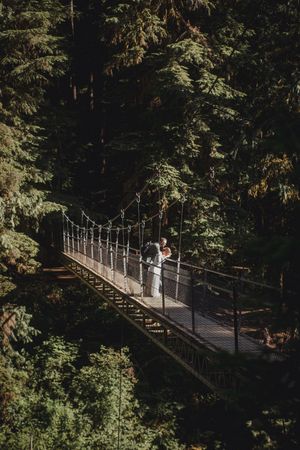 Two figures stand together on a scenic suspension footbridge surrounded by lush evergreen forest.