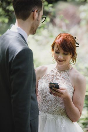 A couple in wedding attire stands together outdoors while the bride looks at a phone in her lace dress.