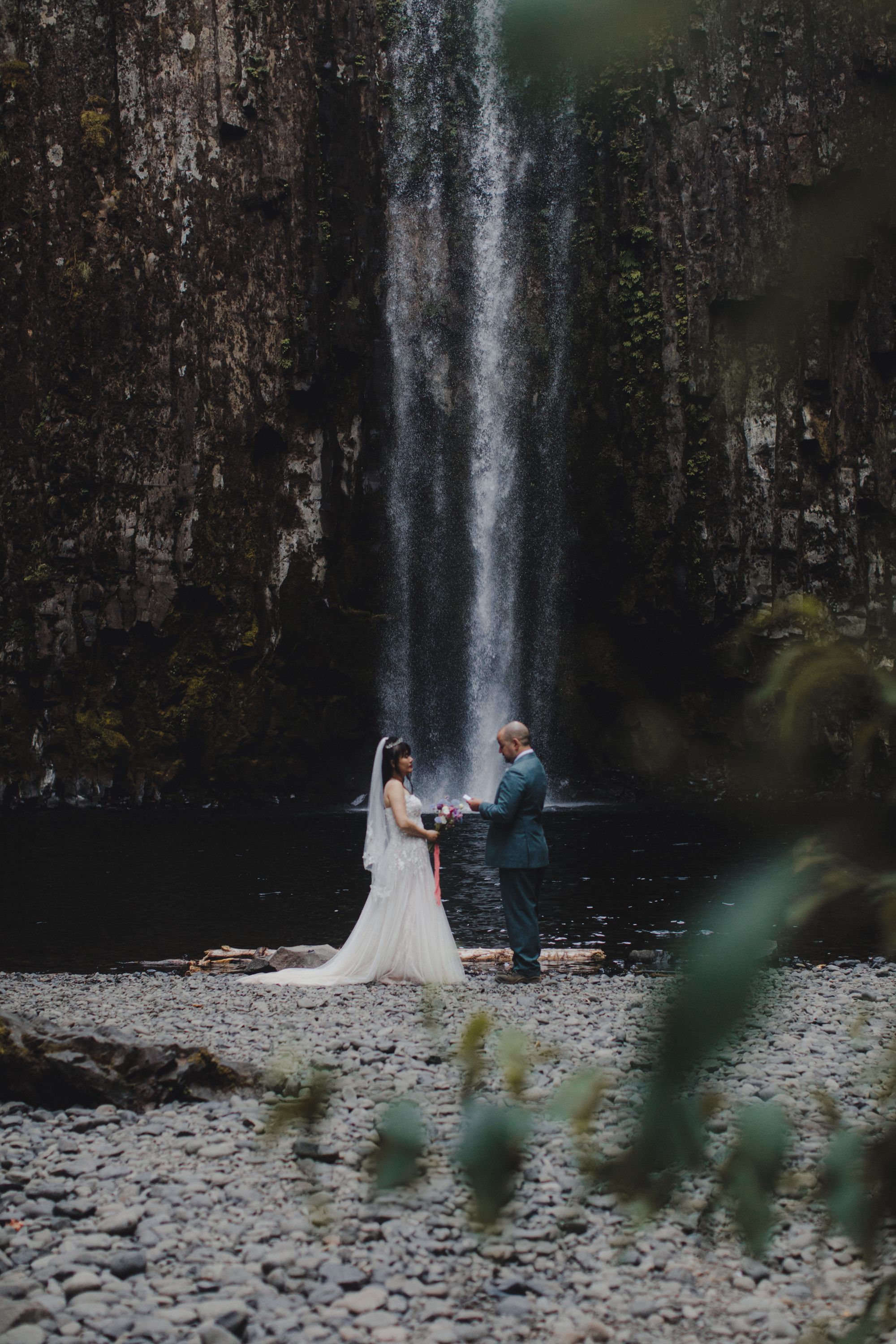 A couple stands on a rocky shore beneath a majestic waterfall in a romantic forest setting.
