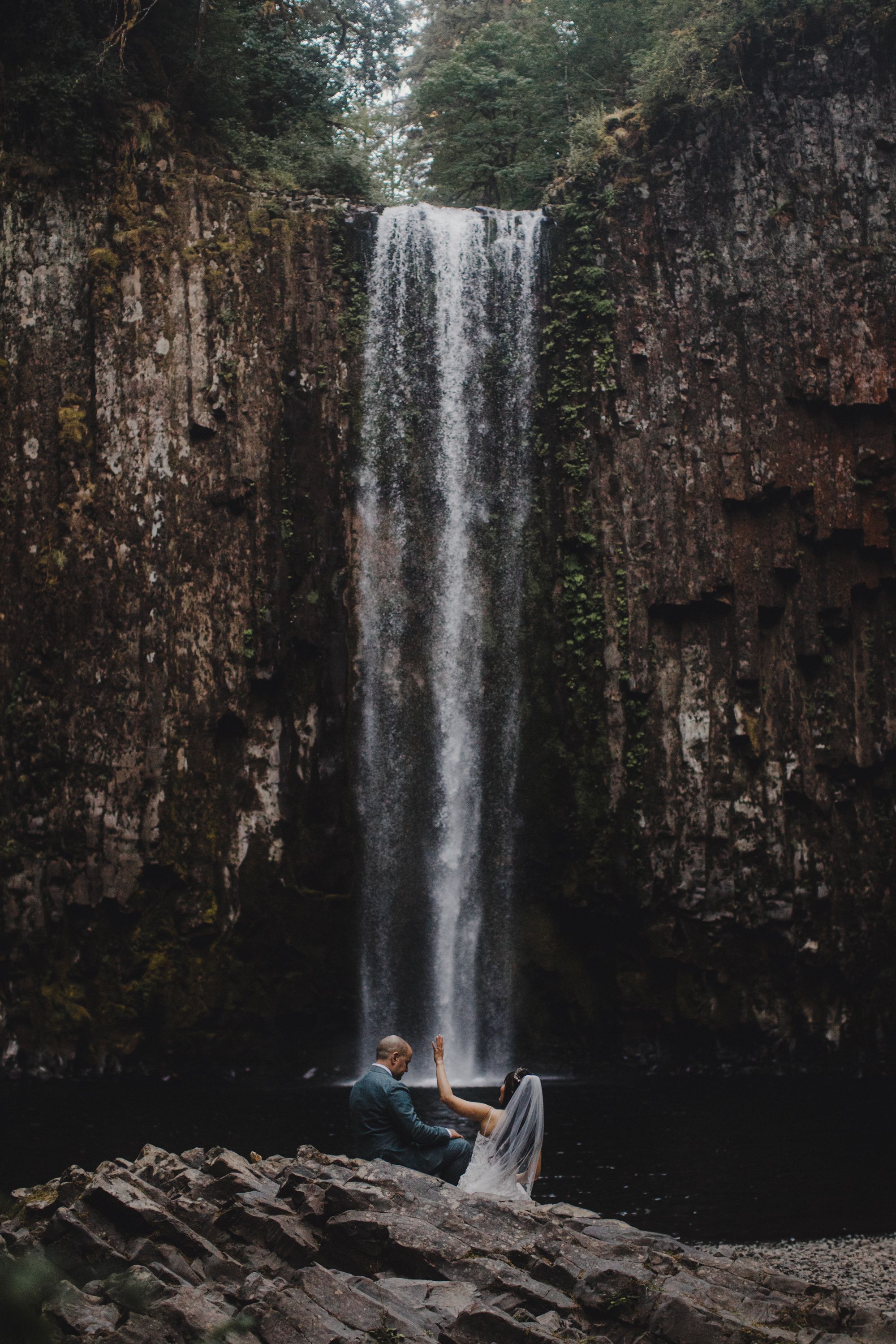 A scenic waterfall cascades down dark rocky cliffs while a person relaxes on stones near the misty pool below.
