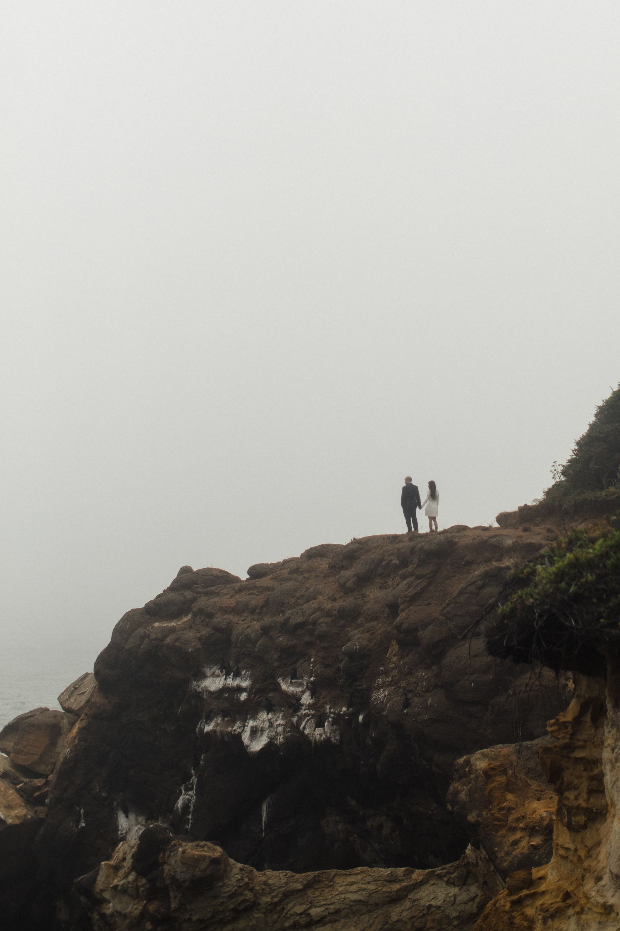 People silhouetted against foggy sky while standing on cliffs watching ocean waves below at sunset.
