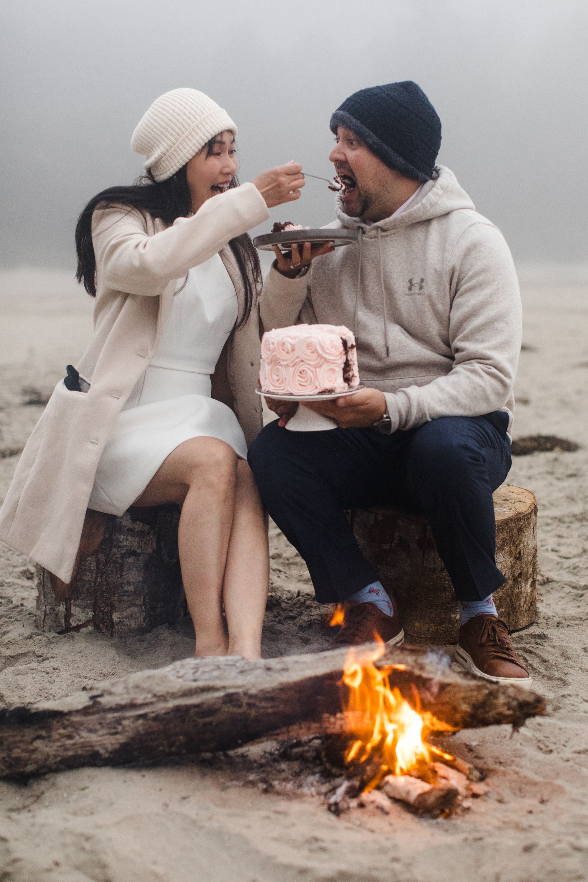 Cozy couple enjoying hot drinks and sharing food by a campfire on a misty beach.