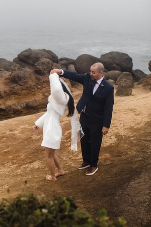 Couple dancing on a scenic coastal cliff with large rocks and misty ocean in the background.