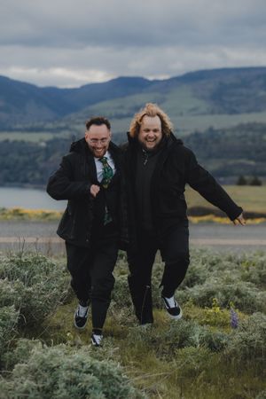 A fun loving couple runs and jumps together on a scenic mountainside meadow covered in sagebrush and wildflowers.