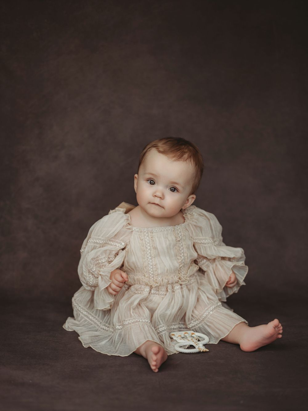 A baby in a vintage white lace dress sits on a dark gray backdrop in a series of artistic portrait poses.