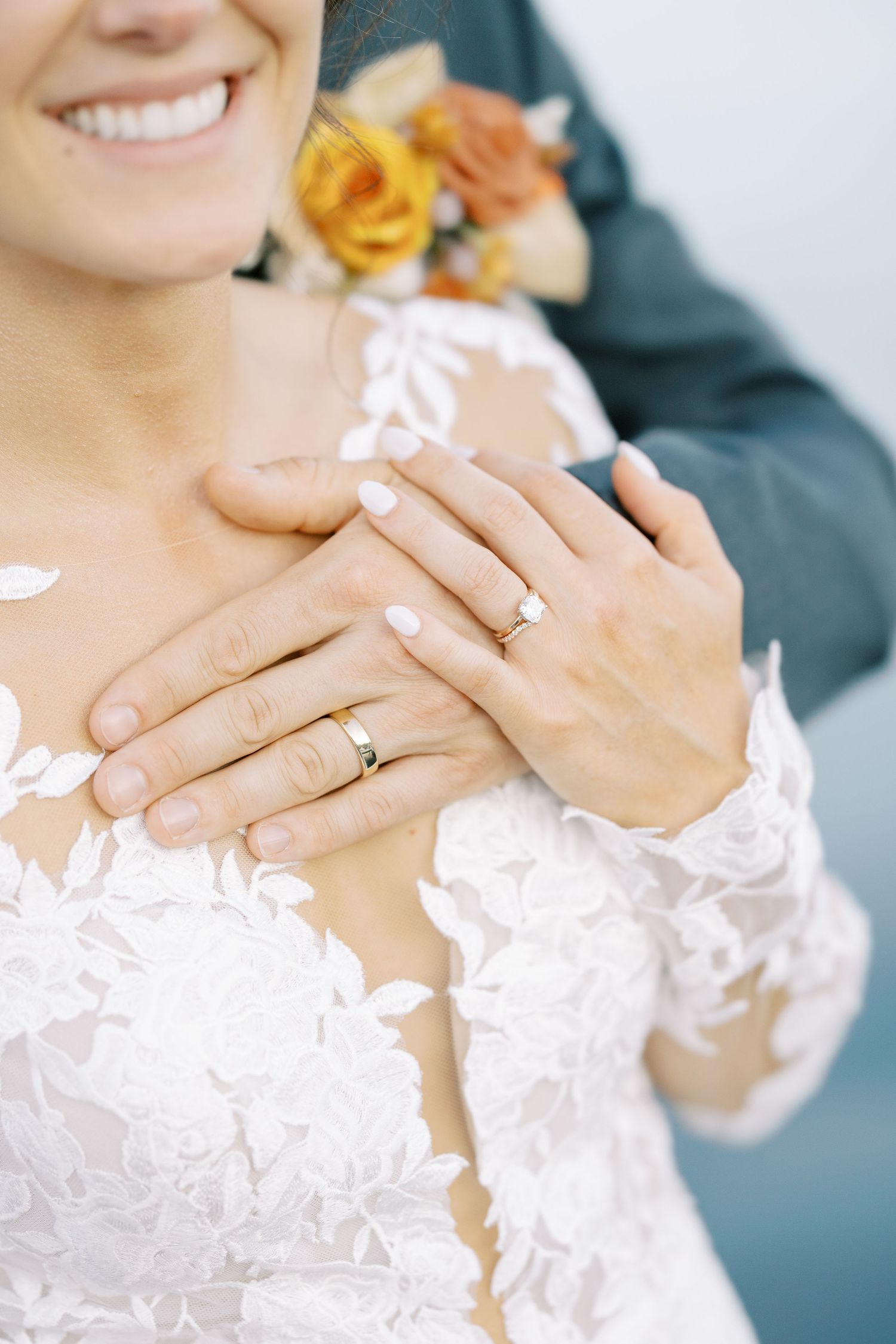 Close-up of wedding rings and delicate lace dress detail with peach and yellow roses in the background.
