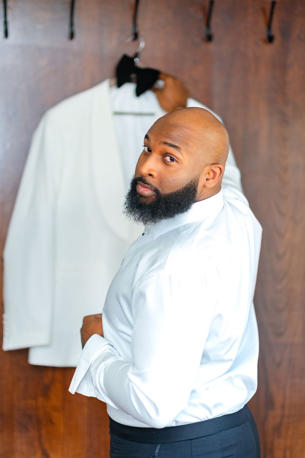 Man in white dress shirt and dark pants poses against wooden wall with clothing hanging in background.