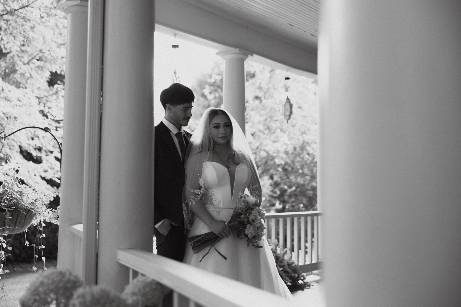 A couple shares a tender moment on a porch with white columns during what appears to be a wedding day.
