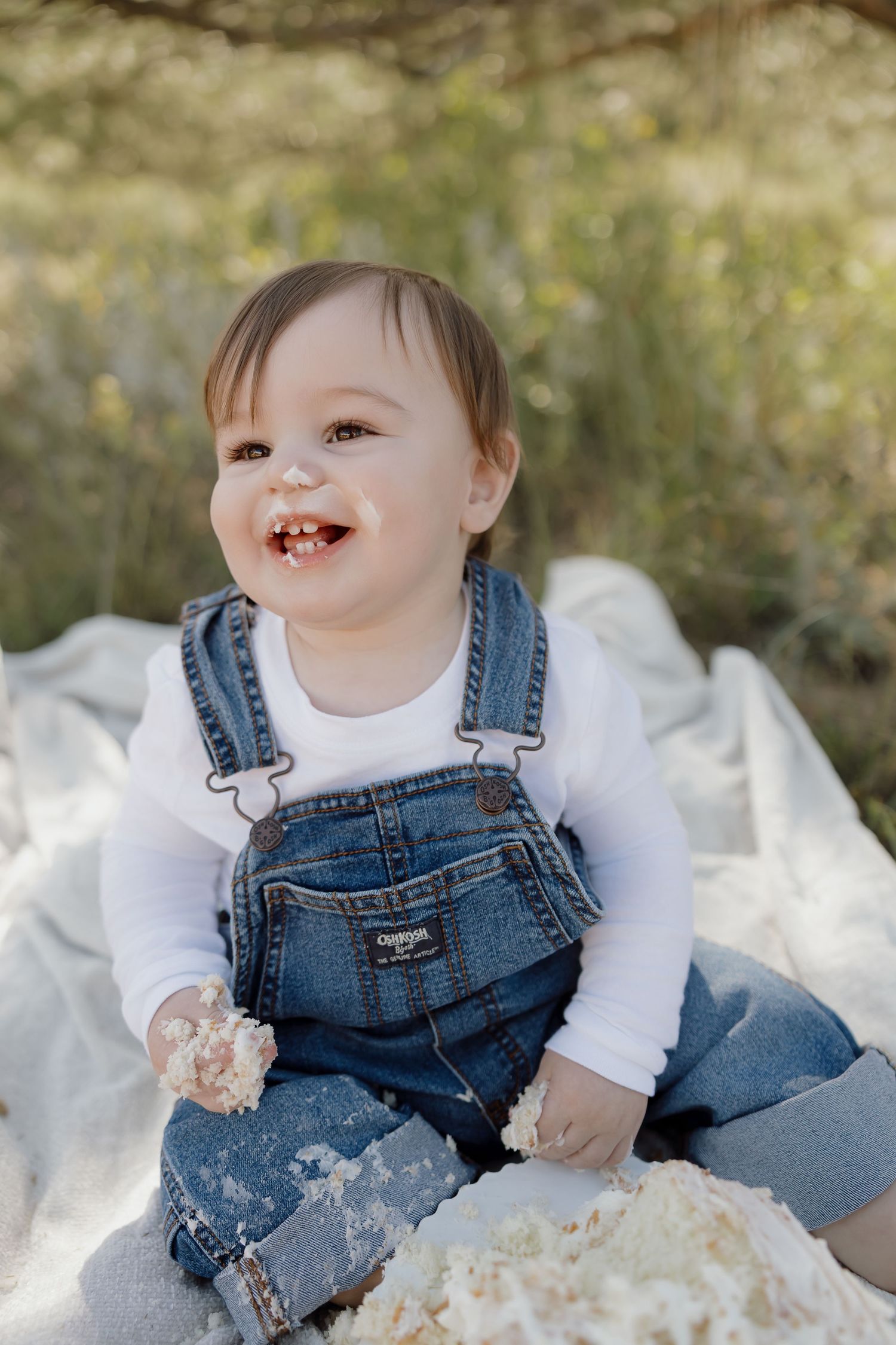 Smiling toddler wearing denim overalls and white shirt sits on blanket outdoors in natural lighting.