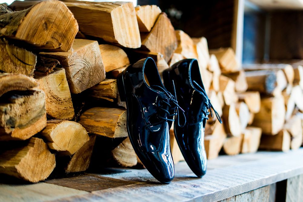 Black dress shoes sit on a wooden shelf against a backdrop of stacked firewood logs.