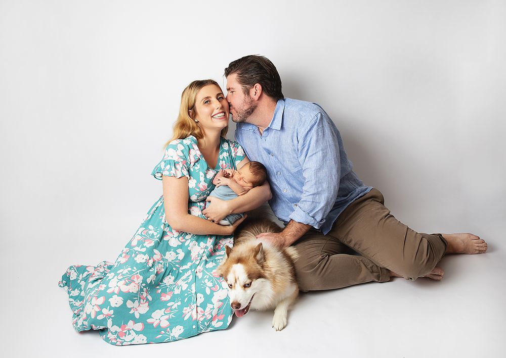 A couple in casual attire lies on the floor playing with their golden retriever puppy in a series of sweet moments. During a newborn session