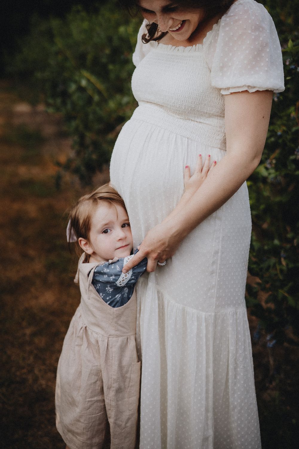 Mother and child share tender moment in white dresses against natural backdrop.
