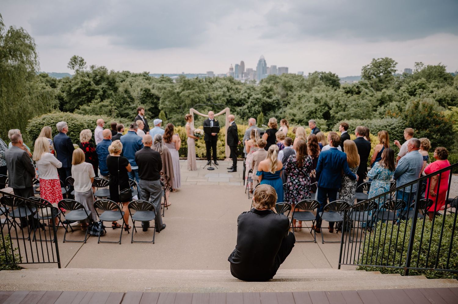 Outdoor wedding ceremony guests seated in colorful chairs overlooking a scenic city skyline on a cloudy day.