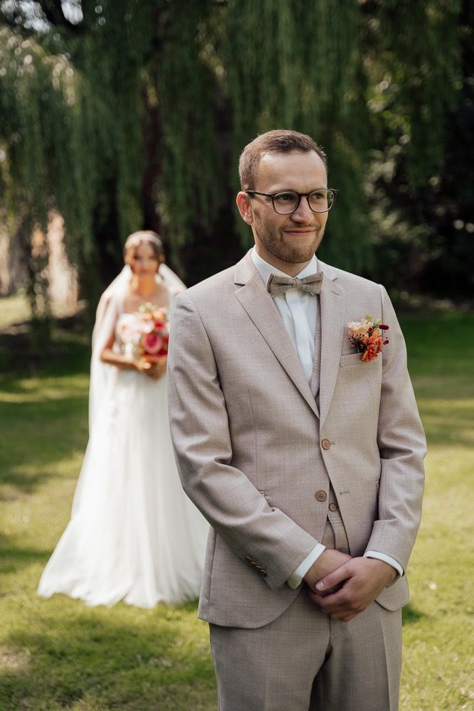 A first look wedding moment captured in a garden setting with a bride and groom in beige suit and white dress.