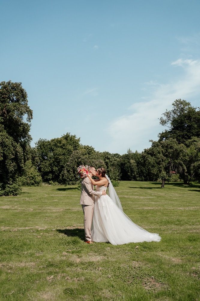 A couple in wedding attire shares a romantic moment together in a sunny green field with trees in the background.