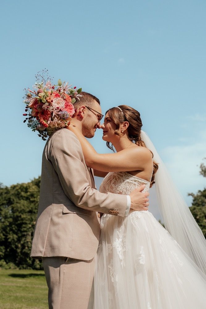 Intimate wedding moment captured against bright blue sky with pink and coral floral arrangements.