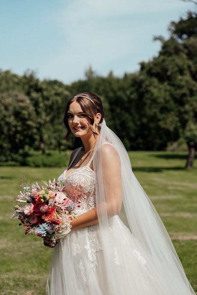 A bride in a flowing white dress and veil holds a vibrant pink and burgundy bouquet in an outdoor garden setting.