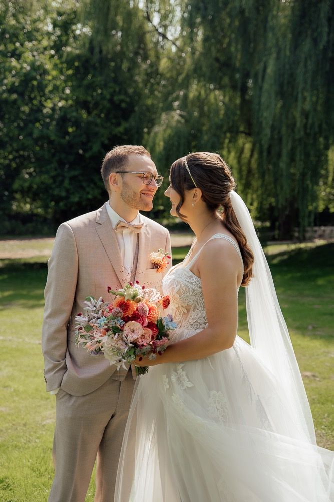 Wedding couple poses in a garden setting with pink and coral floral bouquet and light beige suit.