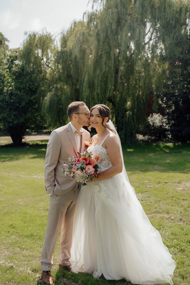 A romantic wedding couple embraces on a sunny day with weeping willow trees creating a dreamy backdrop.