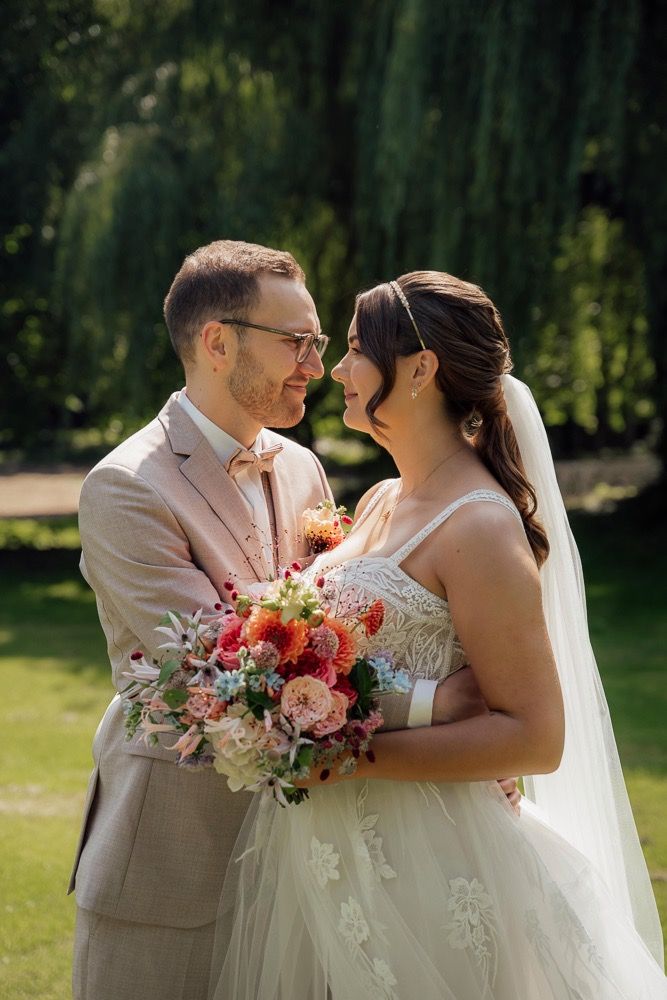 Romantic wedding portraits with couple holding coral and pink bouquet against willow trees.
