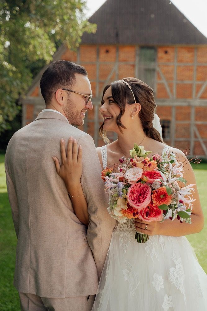 Couple embraces outdoors with bride holding colorful bouquet against rustic building backdrop.