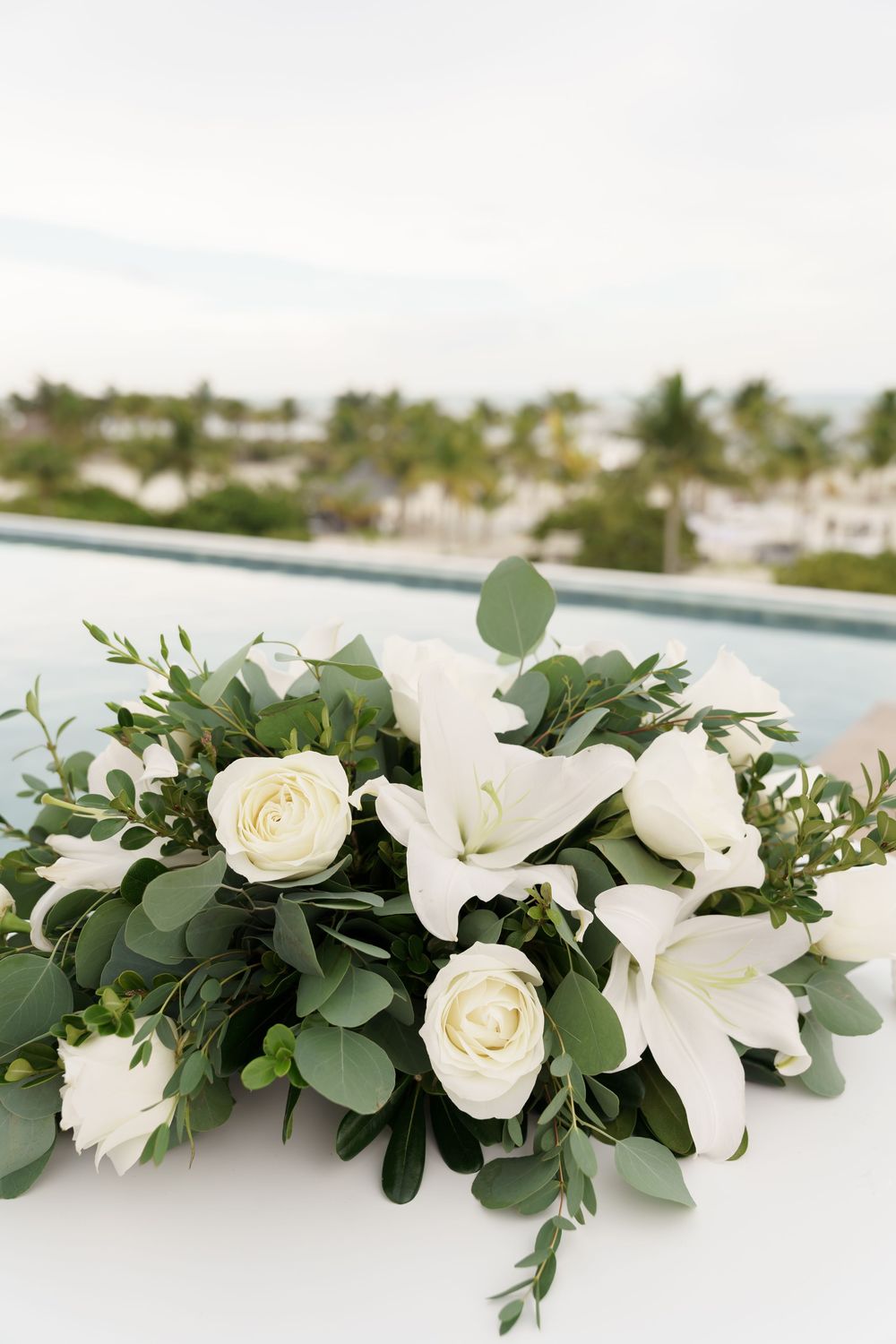 Elegant white floral arrangement with roses, lilies and eucalyptus leaves against a tropical backdrop.