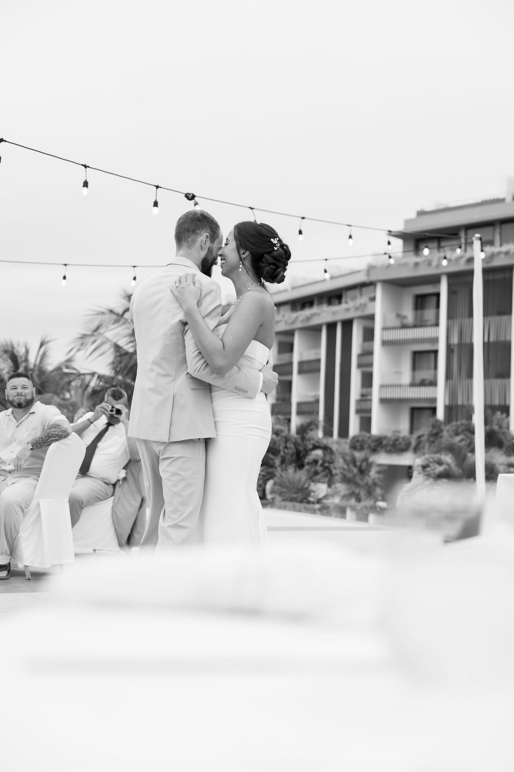 Black and white photo of wedding guests dancing at an outdoor reception with string lights.