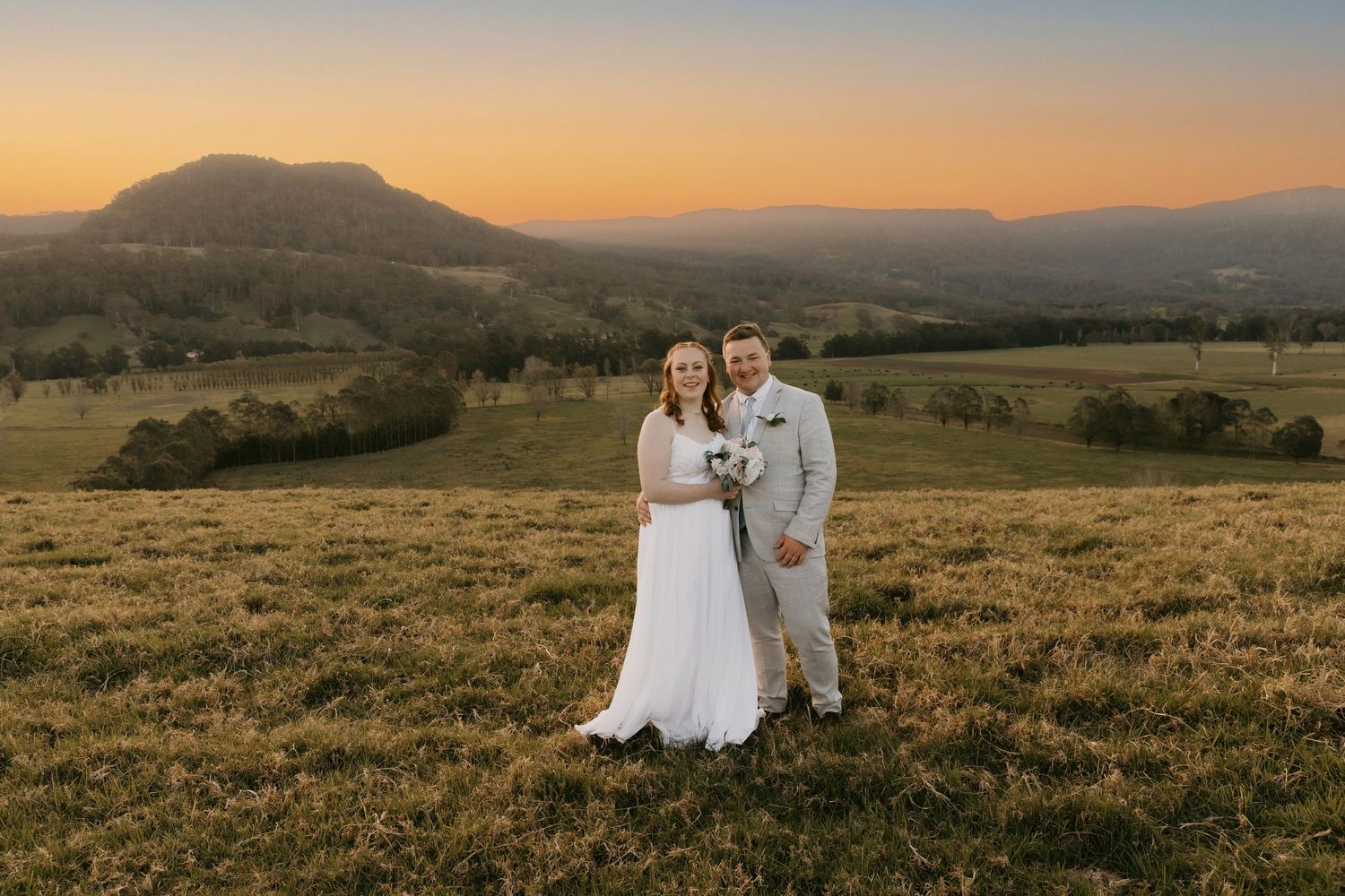 A couple stands in a golden field at sunset with mountains and rolling hills in the background.