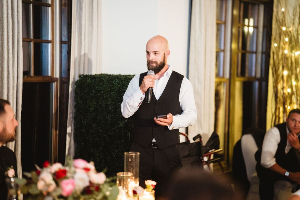 Man in formal attire gives speech at wedding reception with lit candles on dining table.