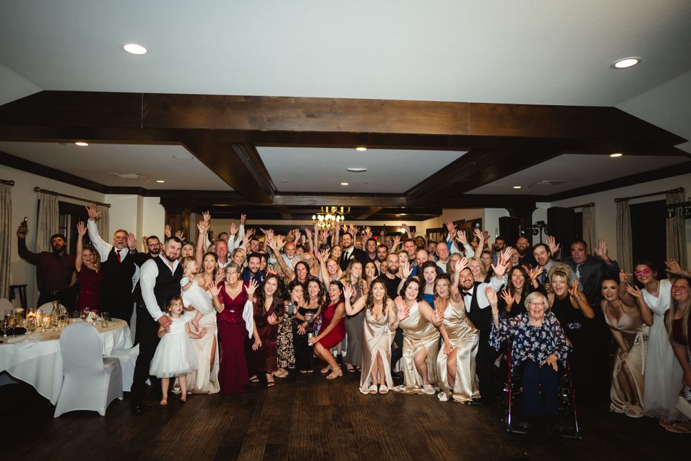 Large group photo of wedding guests posing together in a reception hall with exposed wooden beams.