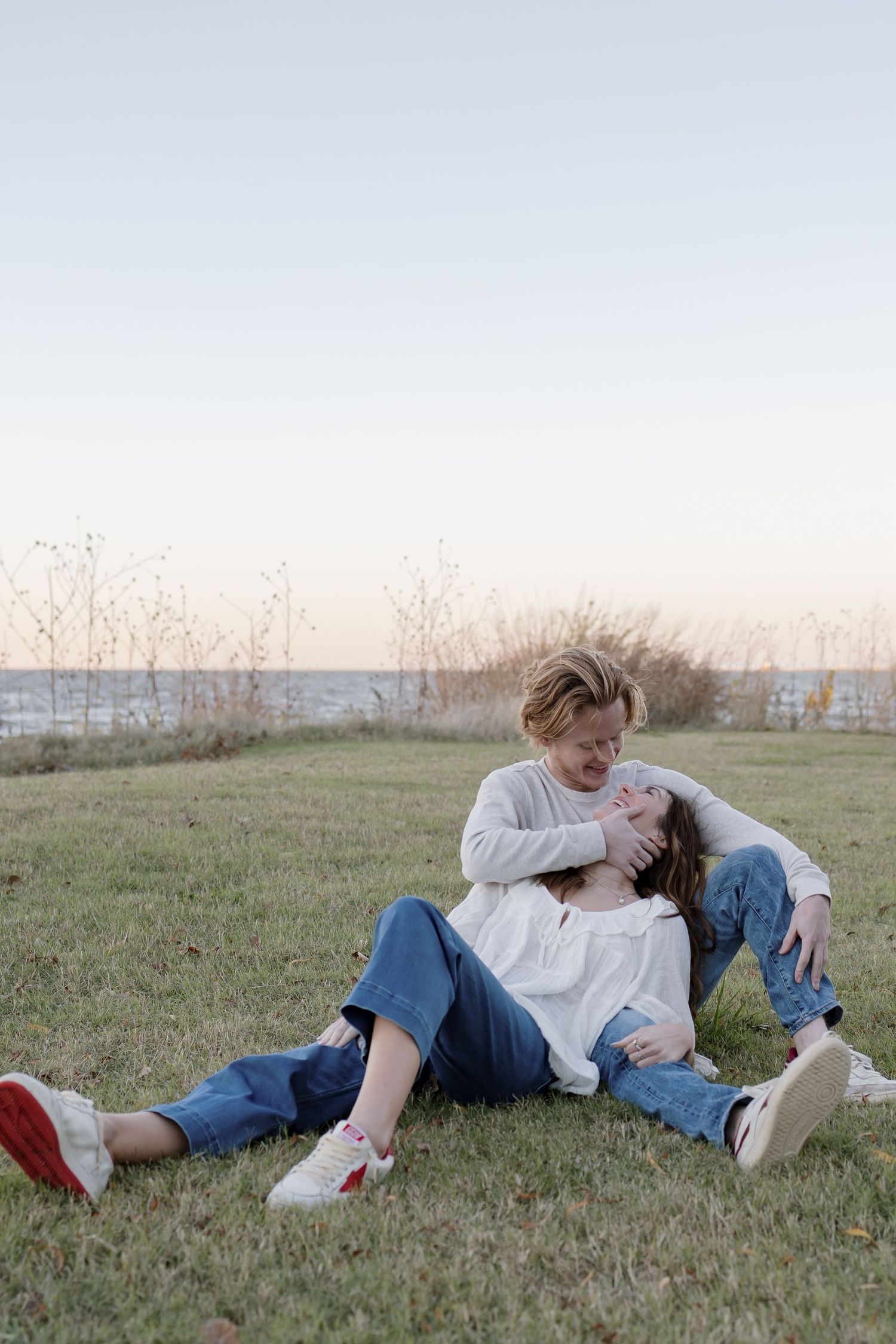 A playful couple in casual attire relaxes together on grass near the ocean.