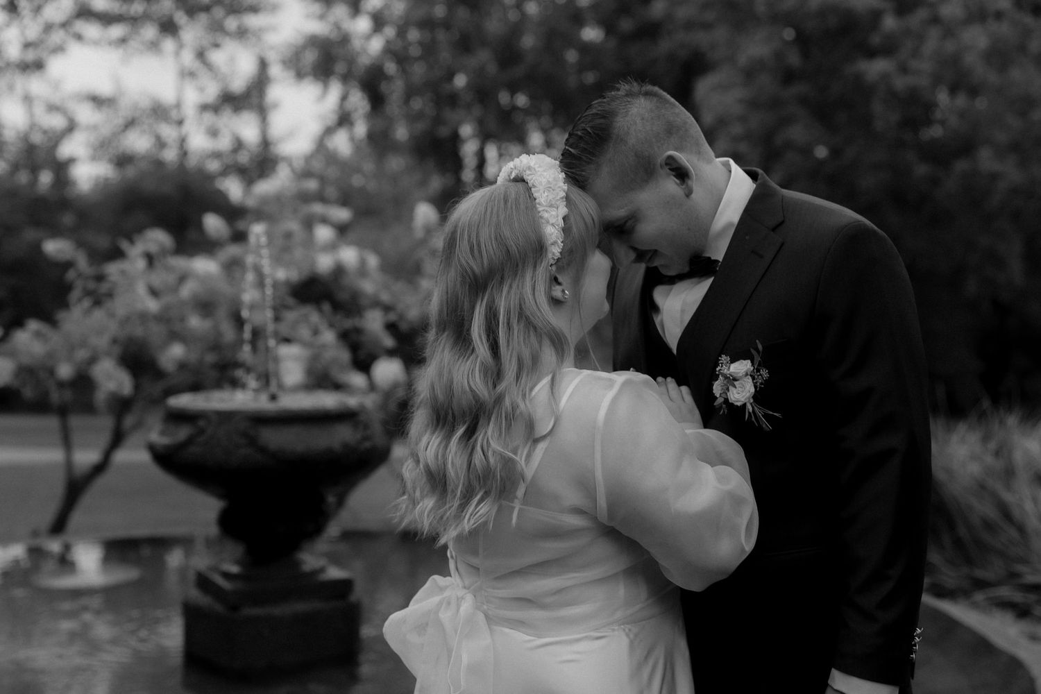 Black and white portrait of bride and groom embracing outdoors near ornate fountain.