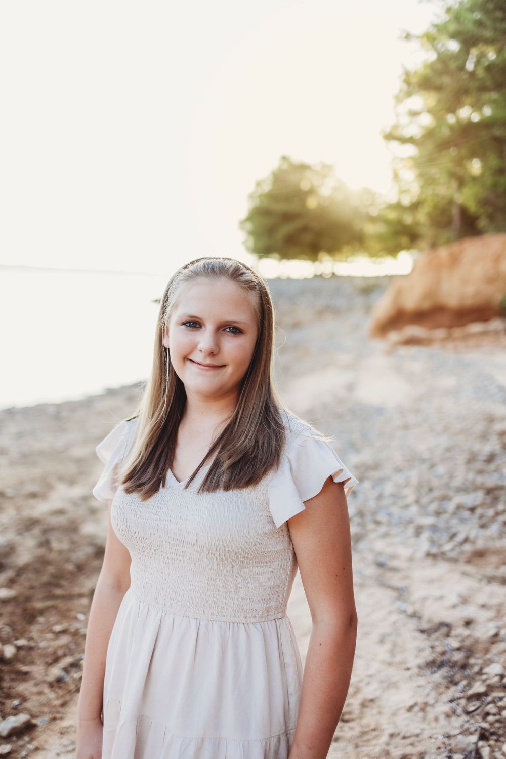 Outdoor portrait of a young person in a white dress standing on a rocky beach at sunset.