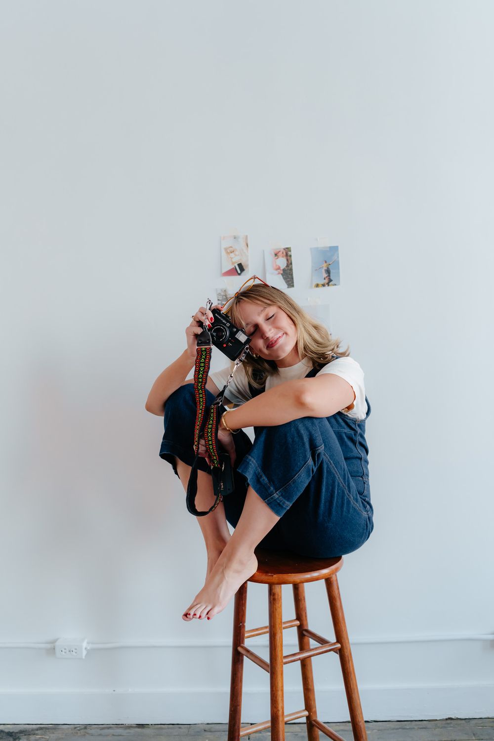 Casual studio portrait of photographer sitting on wooden stool holding camera equipment.