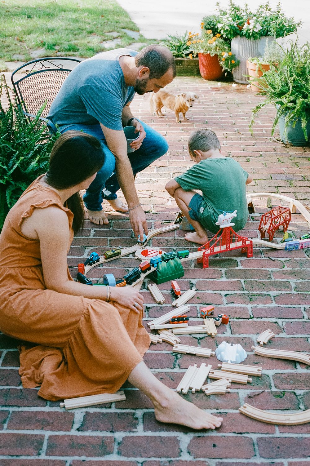Family sitting on brick patio examining toys and tools while cat walks nearby.