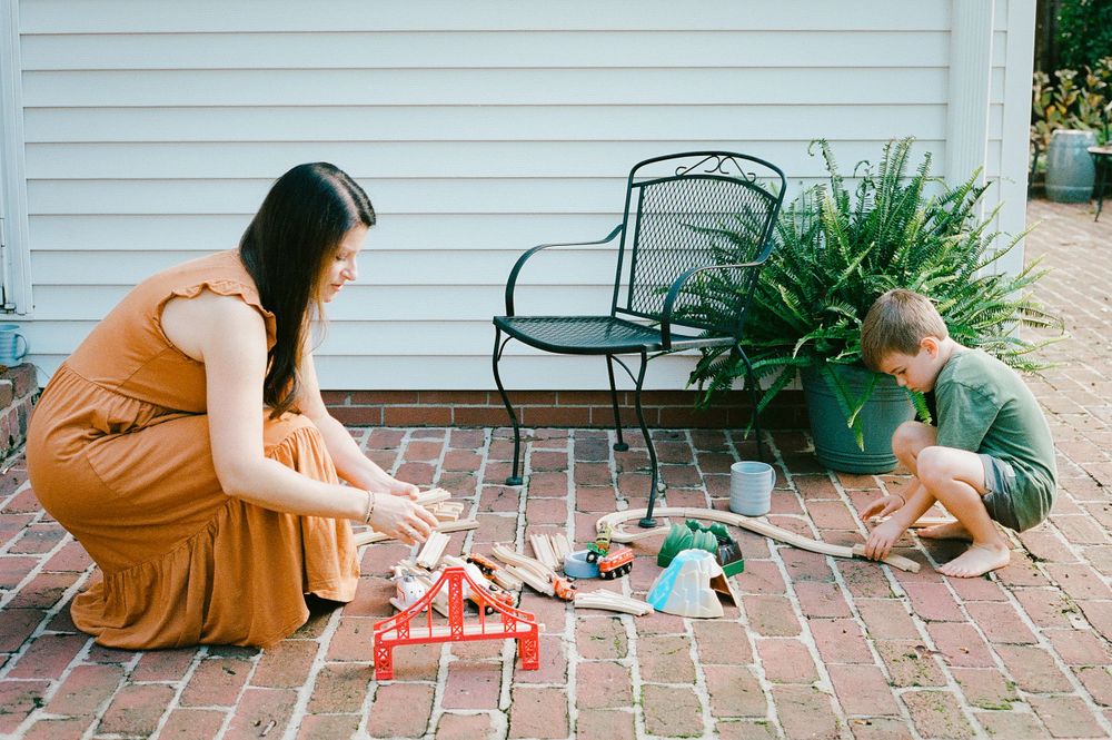 Family playing with toy trains on a brick patio near potted plants and outdoor furniture.